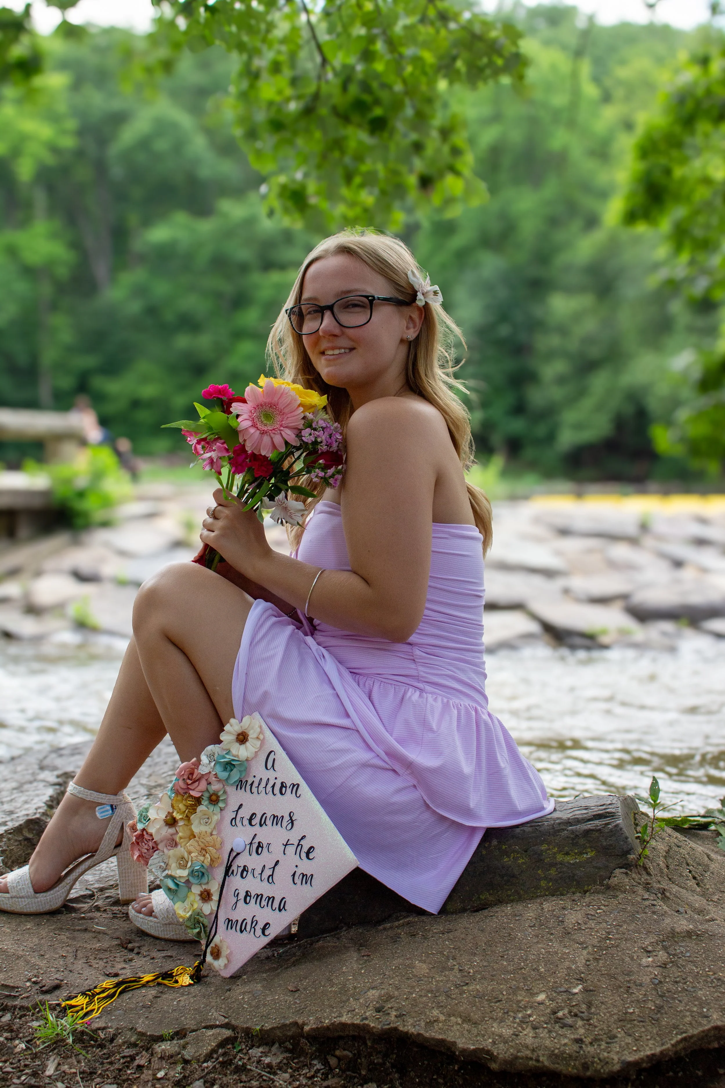 A young woman wearing glasses and a pink dress sitting on a rock by a creek, holding a colorful bouquet of flowers, with a sign that reads 'a million dreams for the world I'm gonna make.' She is smiling and surrounded by green trees in a natural outd