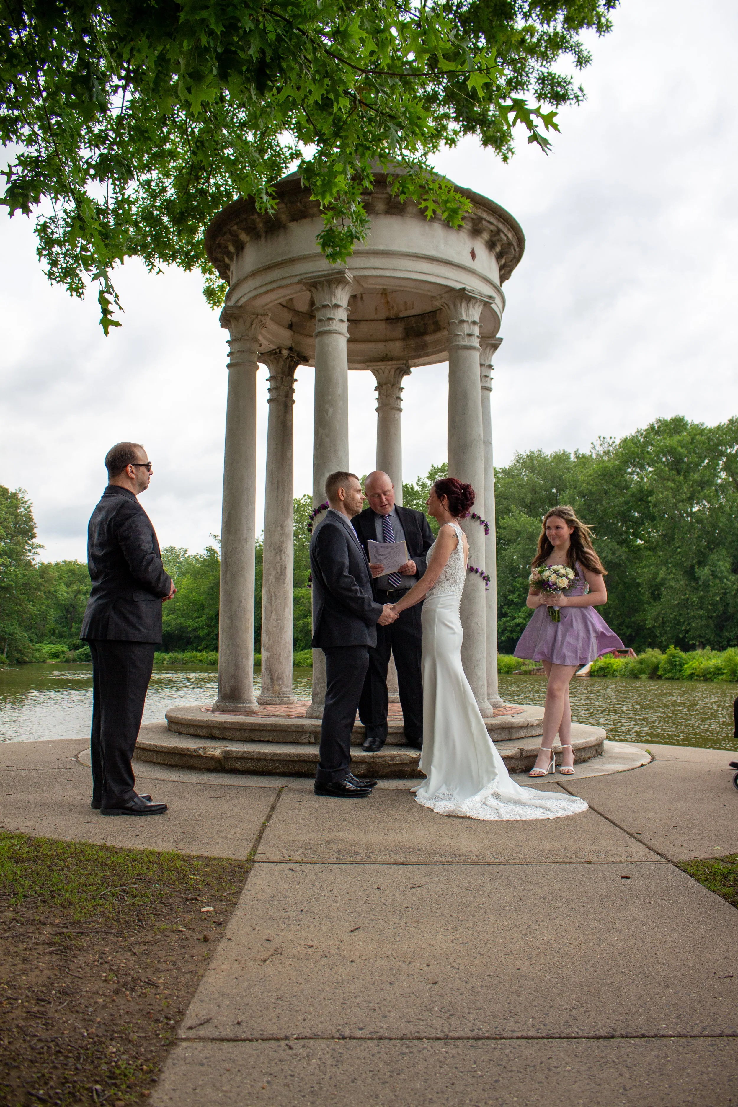 A wedding ceremony taking place outdoors near a body of water, beneath a circular gazebo with stone columns. The bride and groom are holding hands and facing each other, with the officiant standing between them. A bridesmaid holding a bouquet stands 