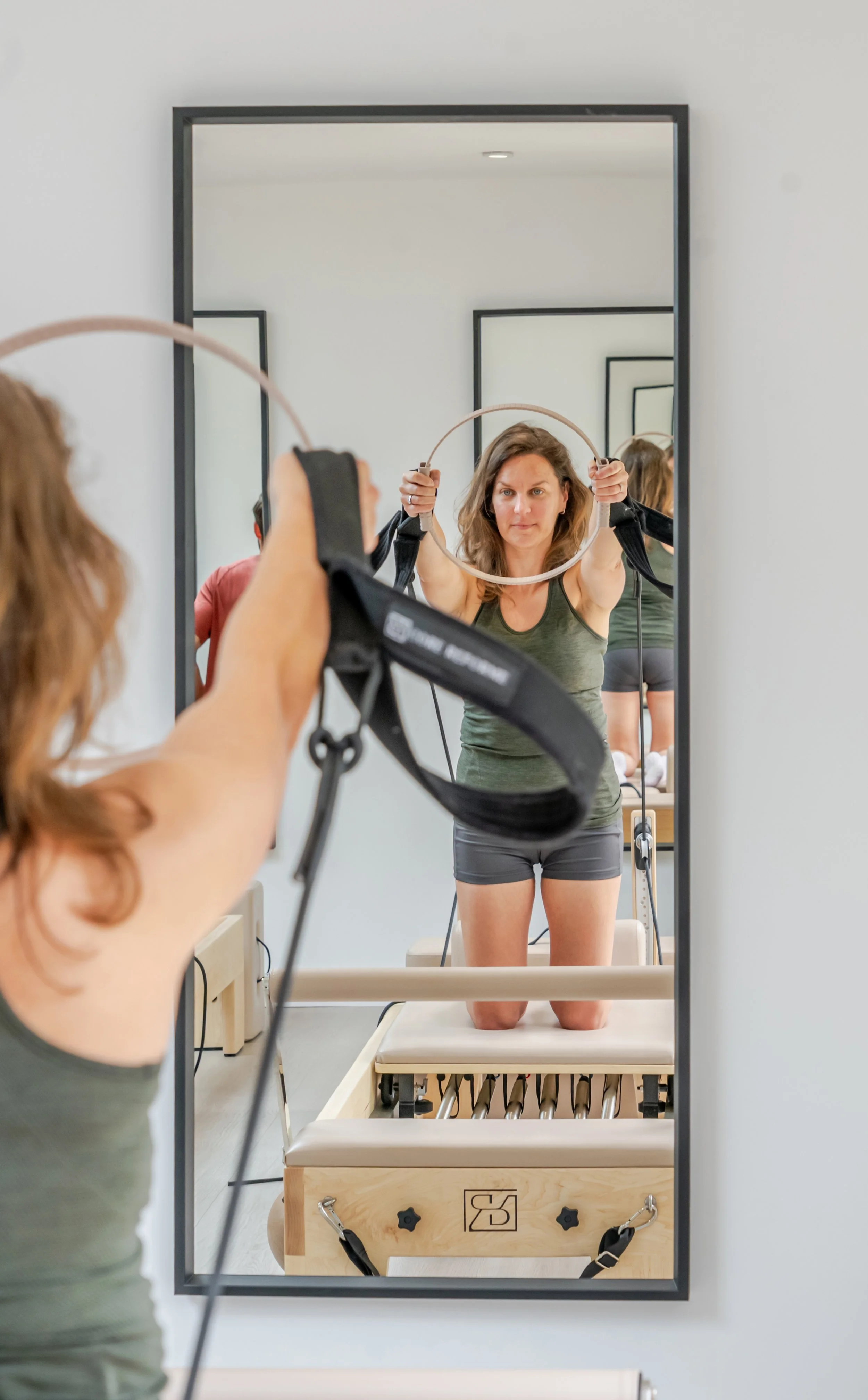Woman doing physical therapy exercises using a Pilates Cadillac machine, seen through a mirror reflecting her kneeling on a padded therapy table.
