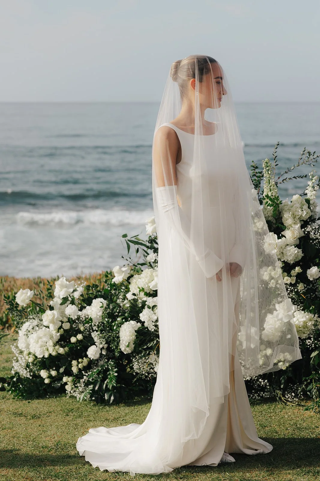 A client of Rachel Lusk Hair and Makeup, a Bridal Hair & Makeup Artist in Napa Valley, offering destination bridal hair & makeup, in a white wedding dress with a veil stands on a grassy area near the ocean, surrounded by white flowers.