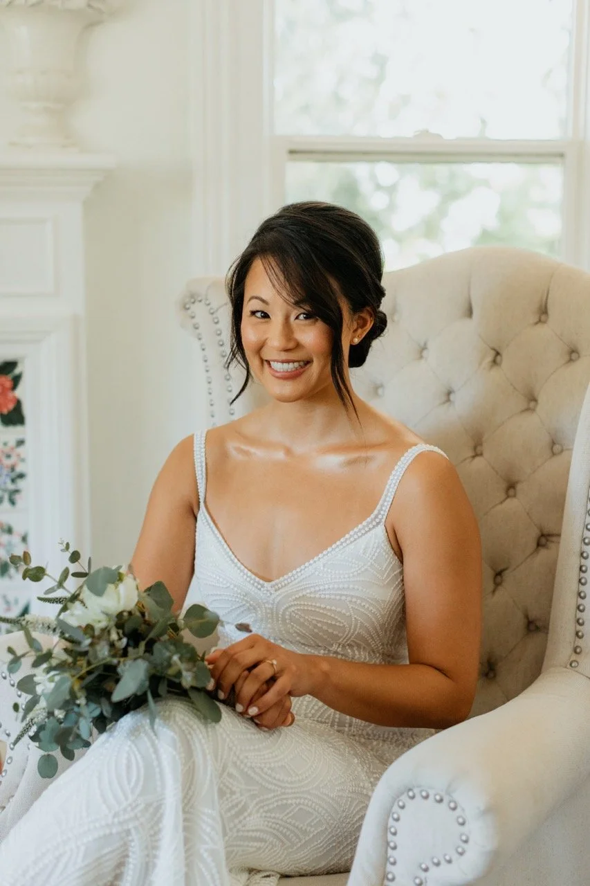 A smiling woman wearing a white dress, sitting on a beige tufted armchair in a bright room with a window in the background. She has dark hair styled up and is holding a bouquet of greenery.