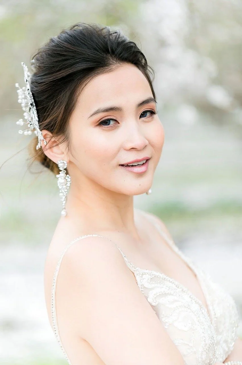 Close-up of a smiling woman with elegant makeup and hair styled in an updo, wearing pearl earrings and a delicate dress with lace details, outdoors with a soft-focus background.