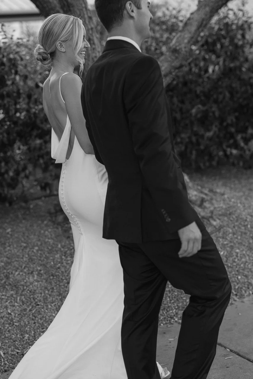 A bride and groom walking outdoors, the bride in a white wedding gown and the groom in a dark suit, both smiling and looking at each other.