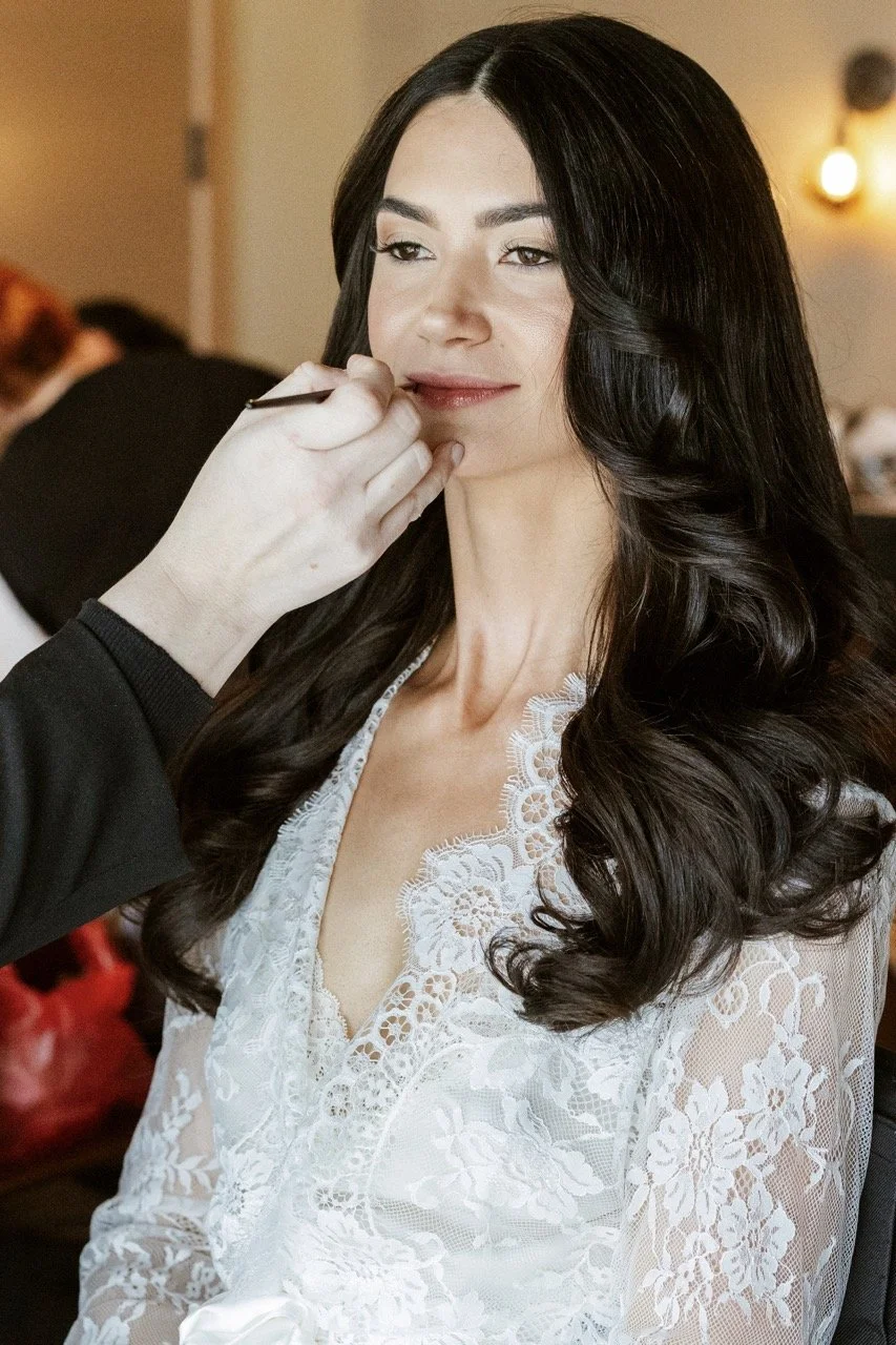 Woman with long, dark, wavy hair getting makeup applied by a makeup artist.