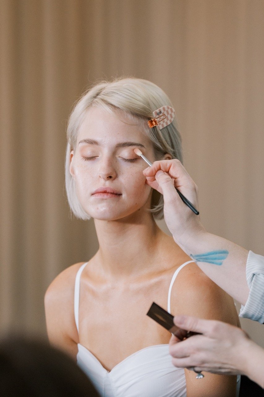 A woman with platinum blonde hair in a white top has makeup applied to her eye area by a person holding a makeup brush.