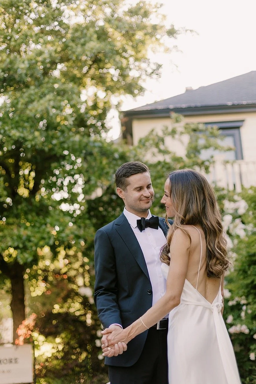A bride and groom holding hands and looking at each other outdoors in a garden, with greenery and a house in the background.