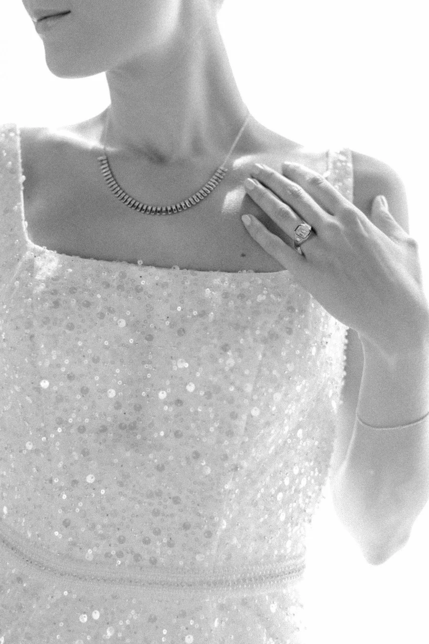 Close-up of a woman wearing a sequined dress, a necklace, and a ring with her hand resting on her shoulder.