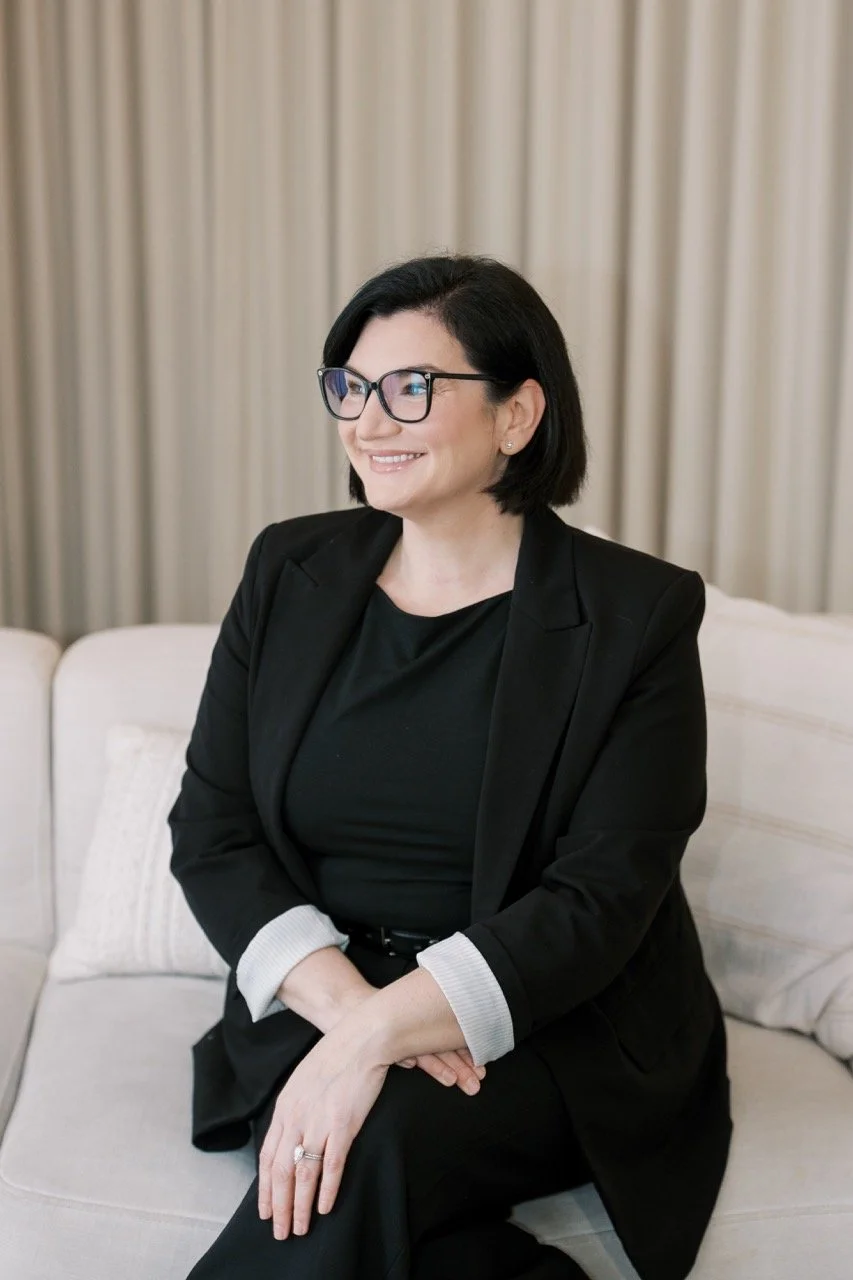 A woman with black hair, glasses, and earrings, wearing a black blazer and shirt, sitting on a beige sofa with a light-colored pillow and beige curtains in the background, smiling and looking to her right.