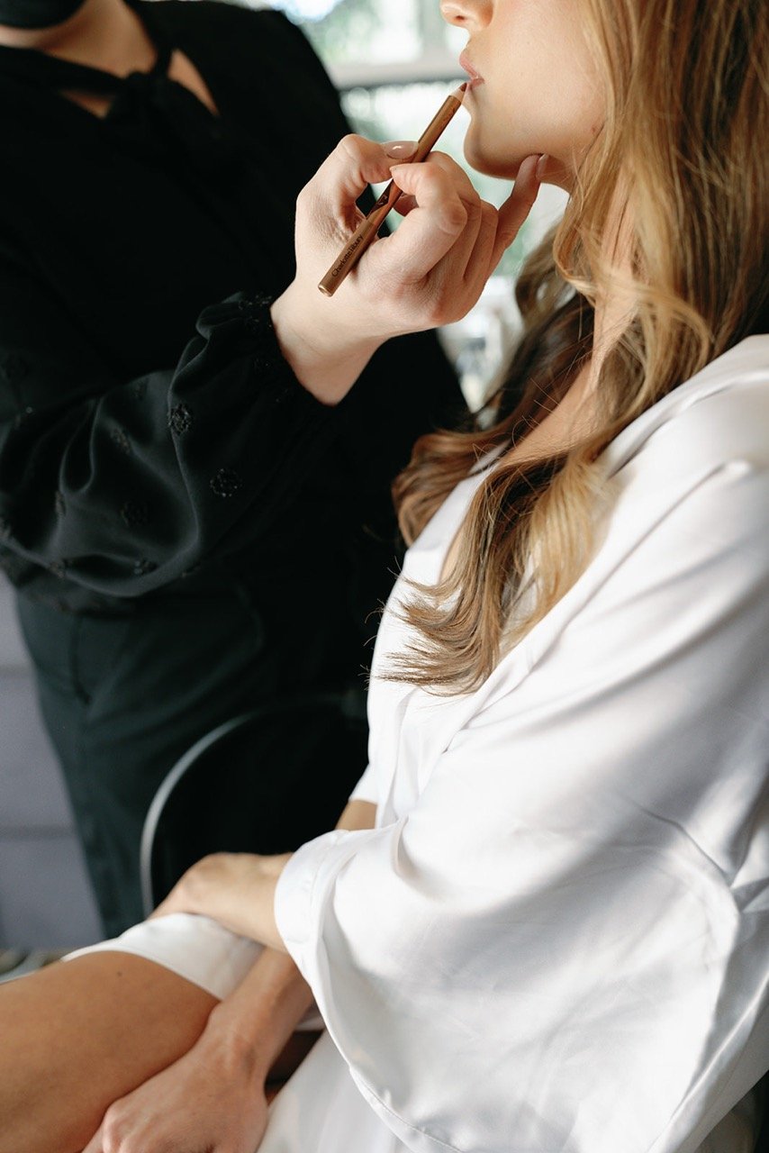 A client of Rachel Lusk, a Napa Valley Bridal Hair & Makeup Artist offering destination bridal hair & makeup, with a makeup artist gently applying makeup near her lips using a brush, while she rests her arm on her lap.