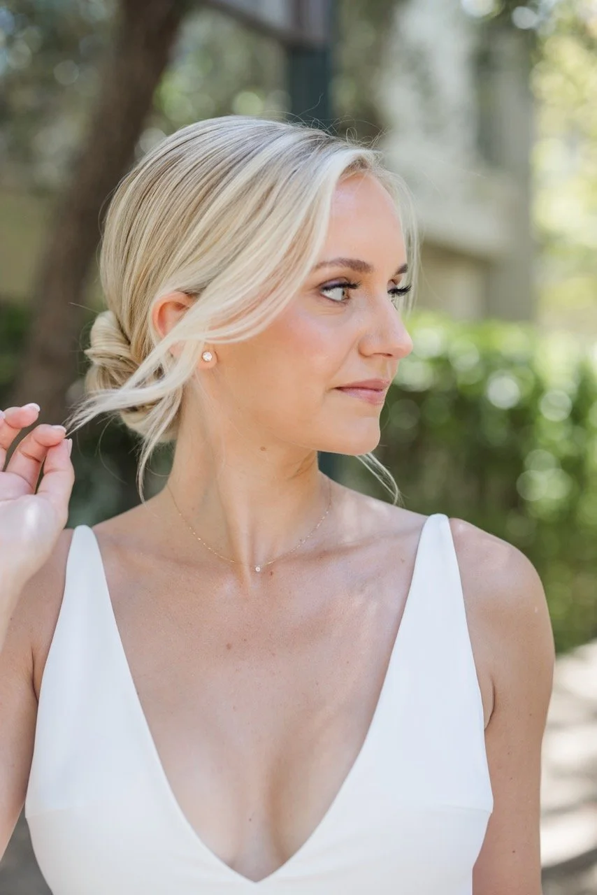 A blonde woman in a white sleeveless dress standing outdoors, adjusting her hair.