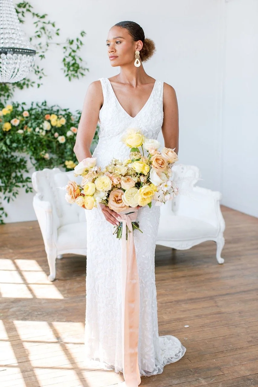 A woman in a white lace dress holding a large bouquet of yellow and peach flowers, standing in a bright room with wooden floors and a white vintage-style sofa.