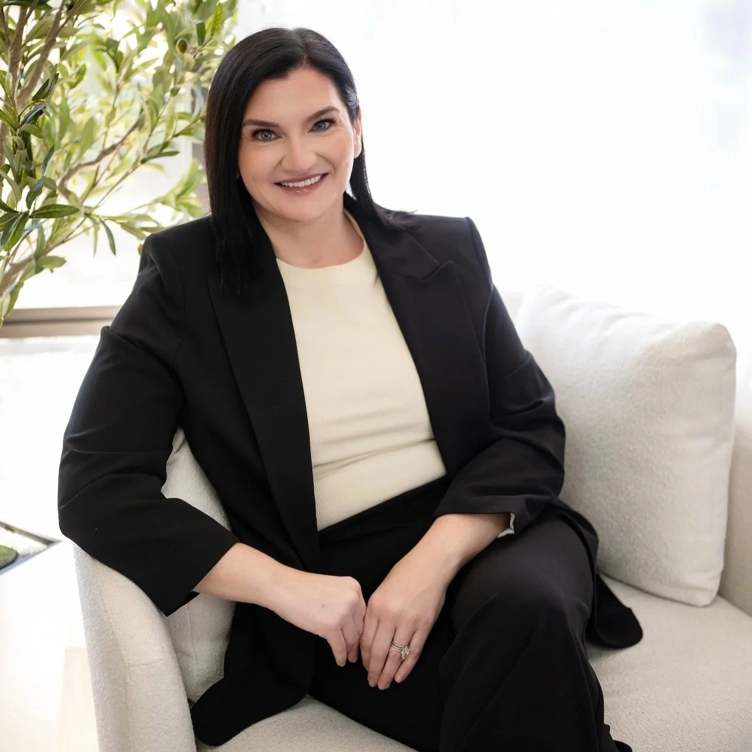 Rachel Lusk, Bridal Hair & Makeup artist offering education for makeup artists wearing a black blazer and cream blouse, sitting on a beige chair with a white pillow. Behind her is a large green plant and a bright window.