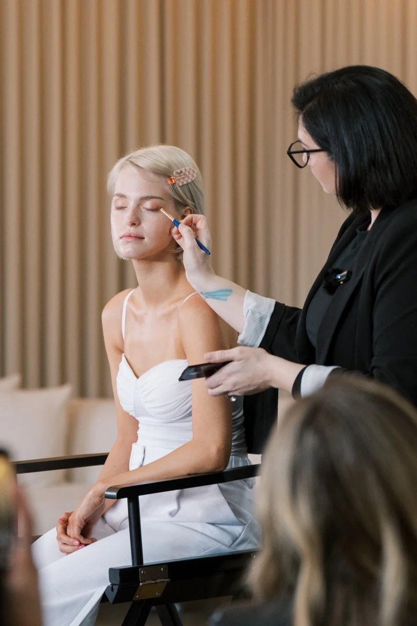 A woman with blond hair is getting her makeup done by a makeup artist with black hair and glasses, in a room with beige curtains.