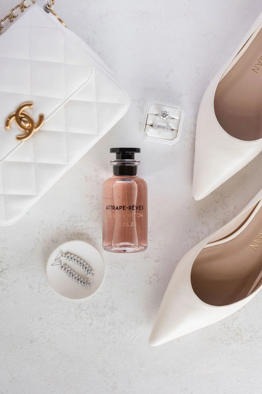 A white Chanel purse, a bottle of Aitrape-Reves perfume, a pair of white high heels, diamond earrings, and rings in a box, arranged on a white surface.