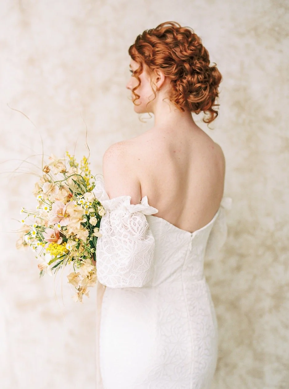A woman with curly red hair in an off-the-shoulder white lace dress holding a bouquet of beige and yellow flowers, standing against a neutral background.