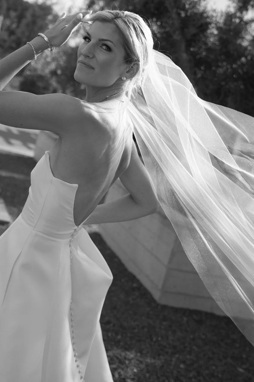 Black and white photo of a woman in a wedding dress with a veil, smiling and holding her hand near her forehead outdoors.