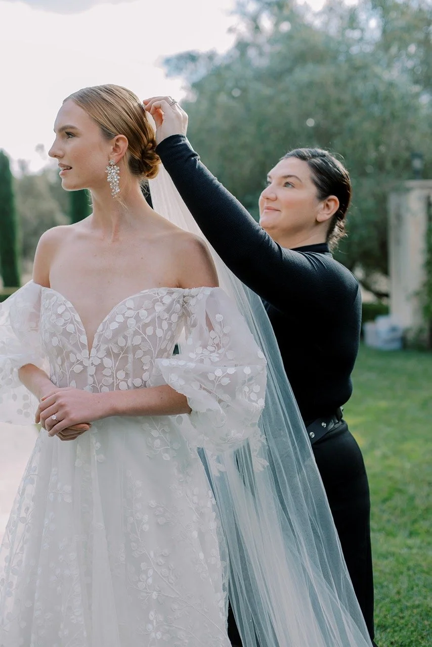 A bride in a white wedding dress with floral embroidery stands outdoors while a person adjusts her veil.