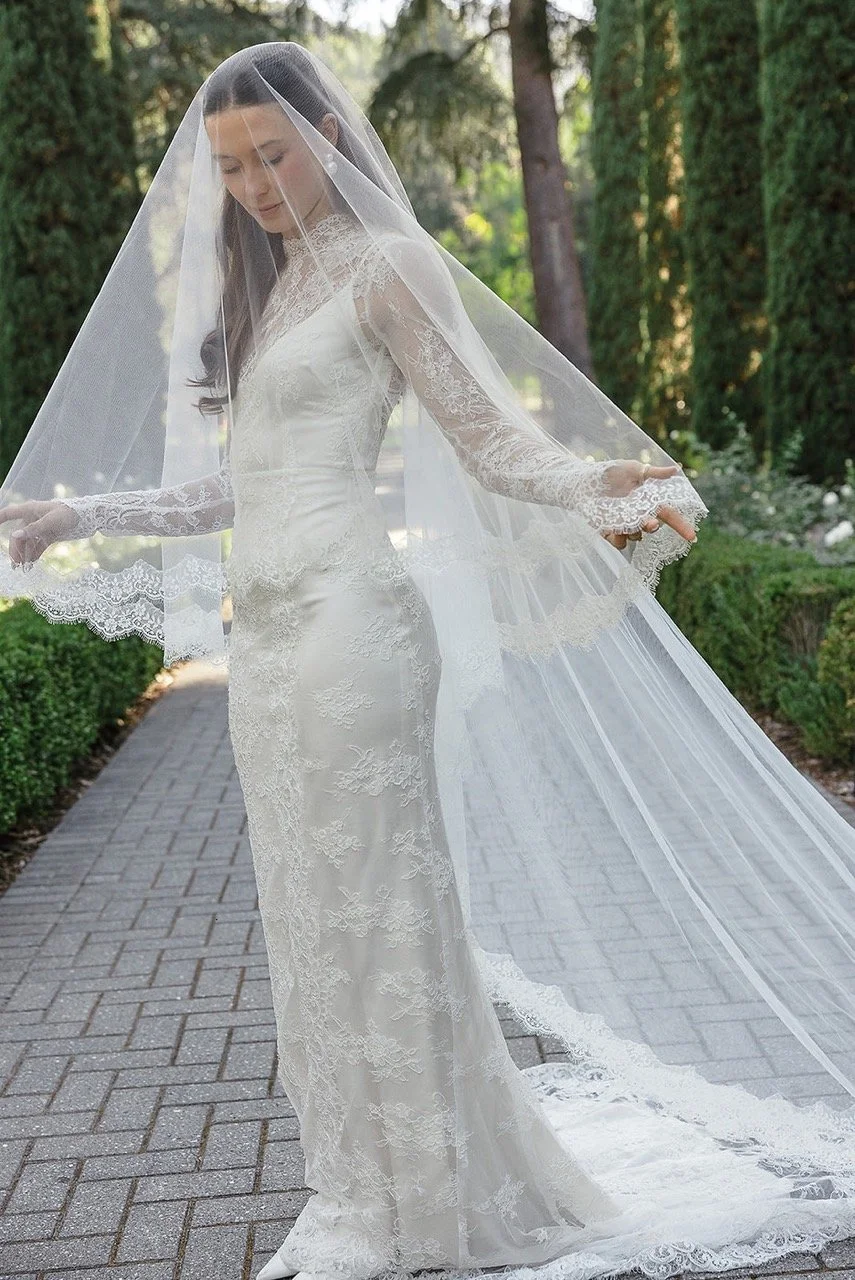 Bride in a long white lace wedding gown and veil walking outdoors on a stone path surrounded by greenery.