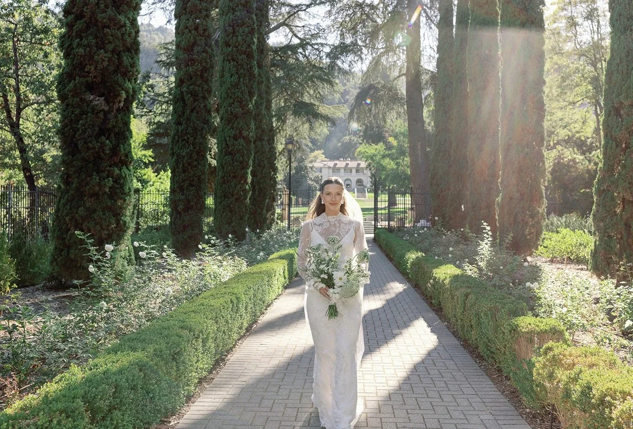 A woman in a white wedding dress holding a bouquet of white flowers walking on a garden path with tall trees and greenery on a sunny day.