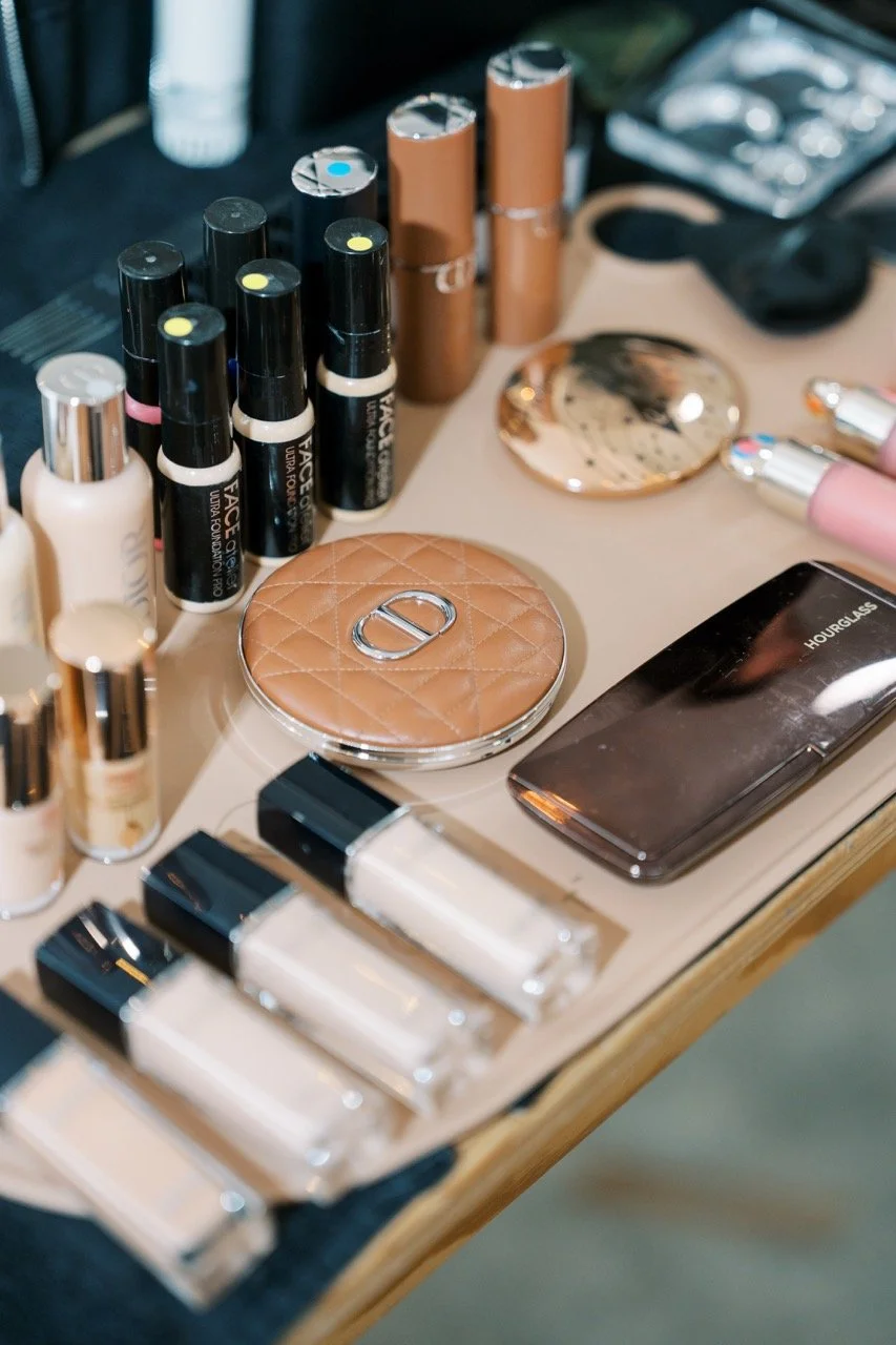 Beauty products on a vanity table including foundation bottles, cream and lip gloss tubes, makeup compacts, and a smartphone with the word 'HOURGLASS' displayed on the screen.