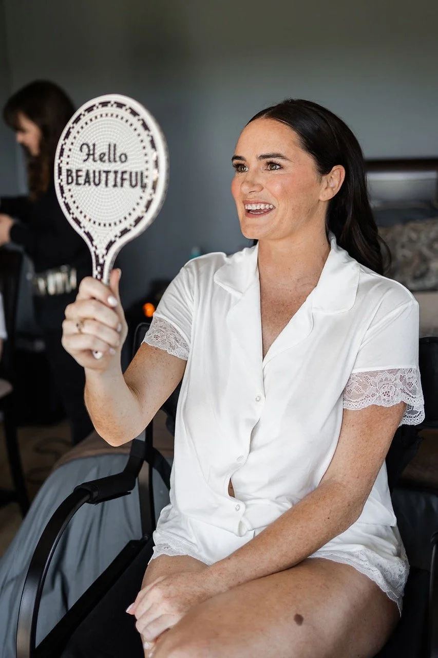 A woman sitting in a wheelchair, dressed in a white satin nightgown, holding a mirror that says 'Hello Beautiful,' smiling, with another woman in the background.