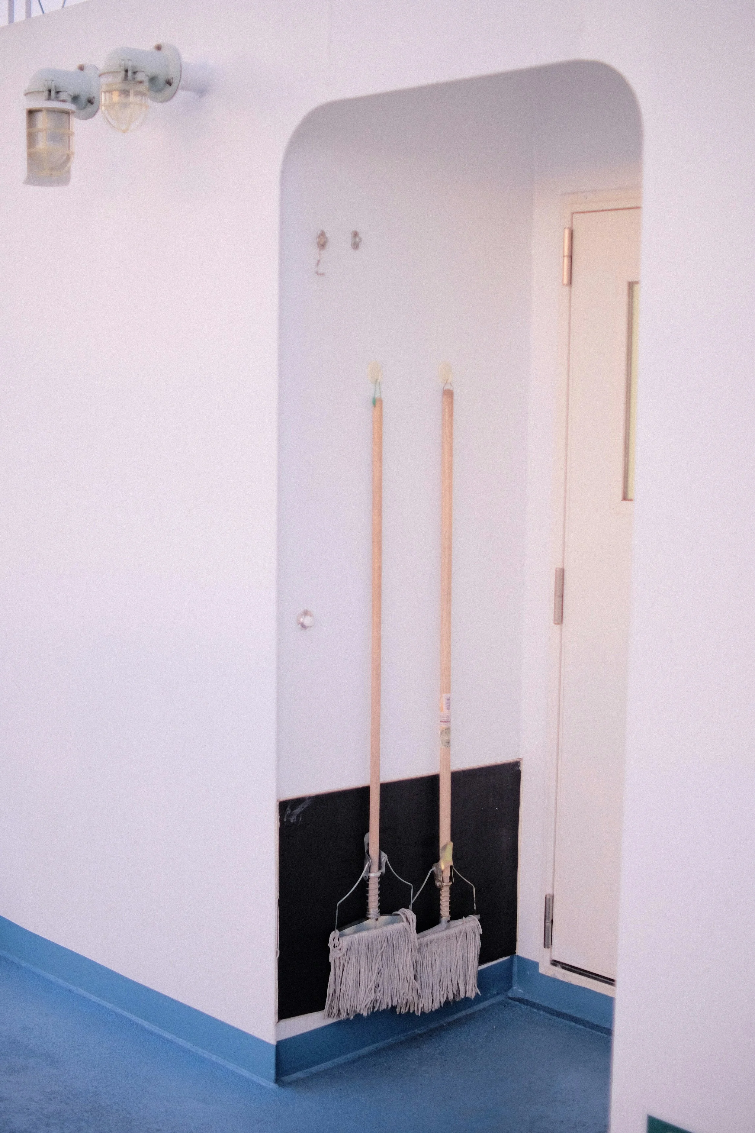 Two mop brooms hanging on a wall with a wall-mounted light fixture and a door in the background.