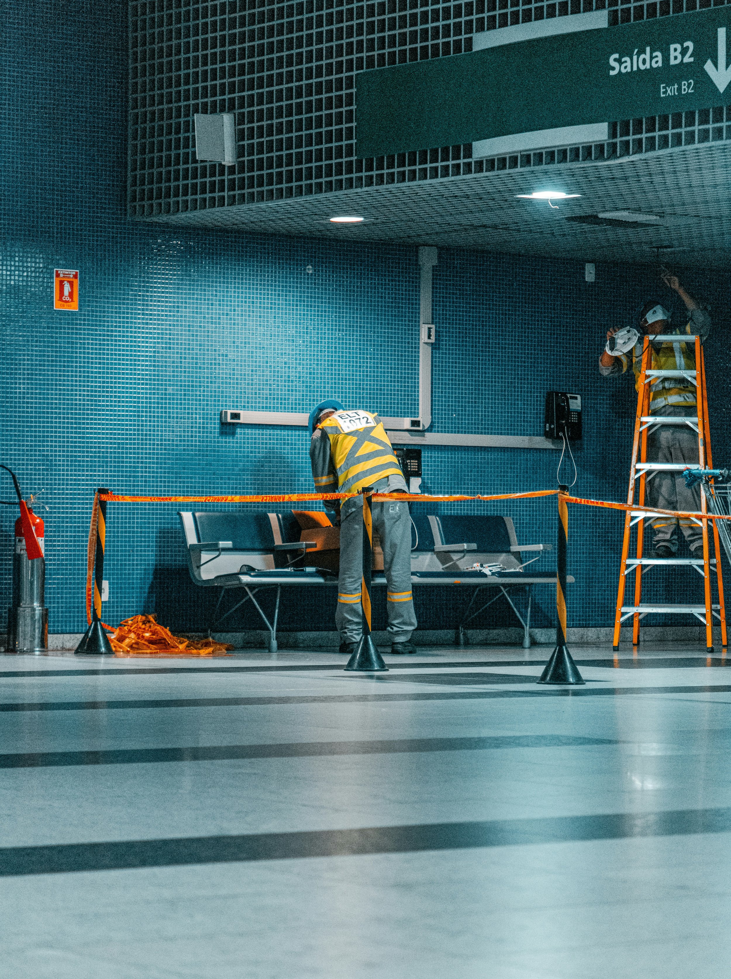 Two maintenance workers wearing yellow safety vests and helmets are working near benches in an airport. One worker is on a ladder, and the other is bending over while working on wiring or panels on the wall. There is a fire extinguisher, orange caution tape, and some equipment on the ground.