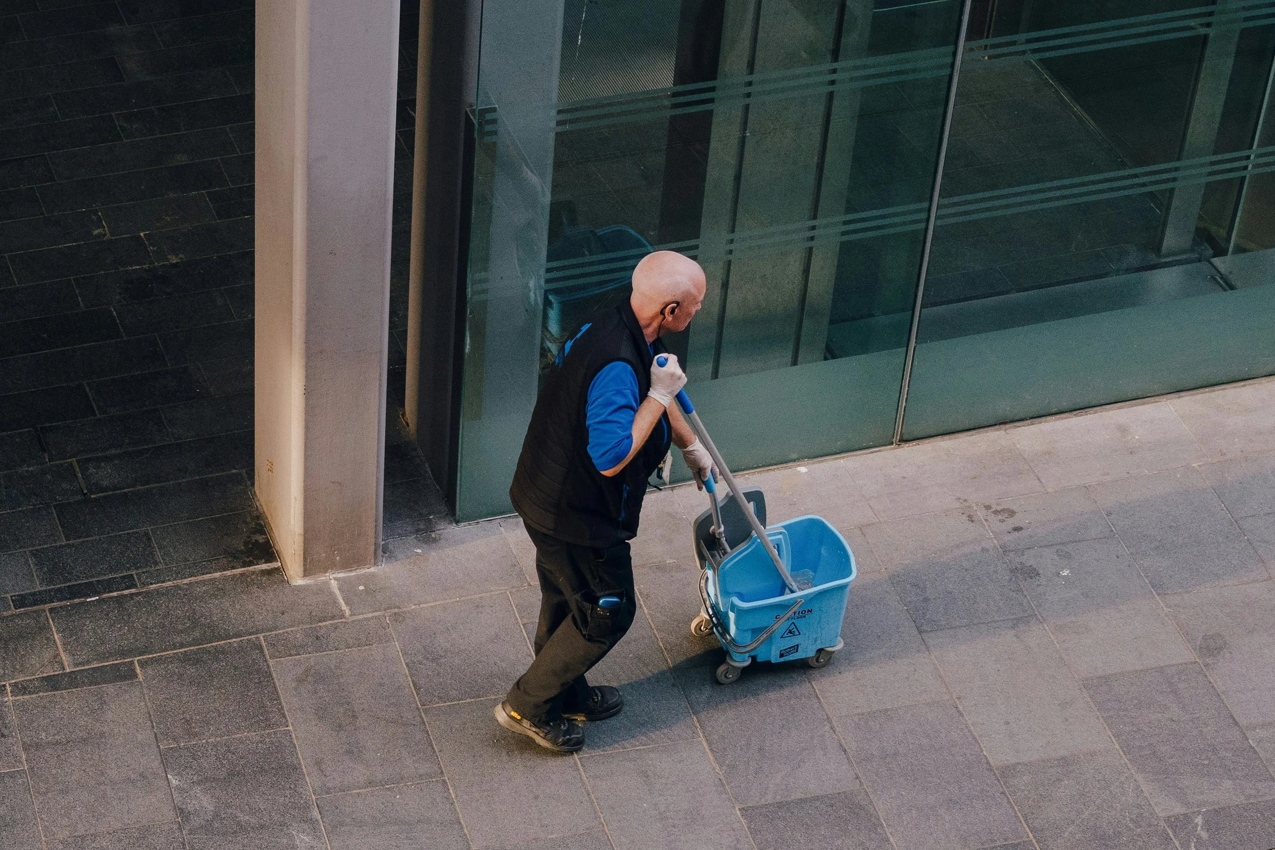 A bald man wearing a black vest over a blue shirt is pushing a blue cleaning cart with a mop inside. He is wearing white gloves and earphones. The scene is in an indoor area with dark tiled floors and glass walls.