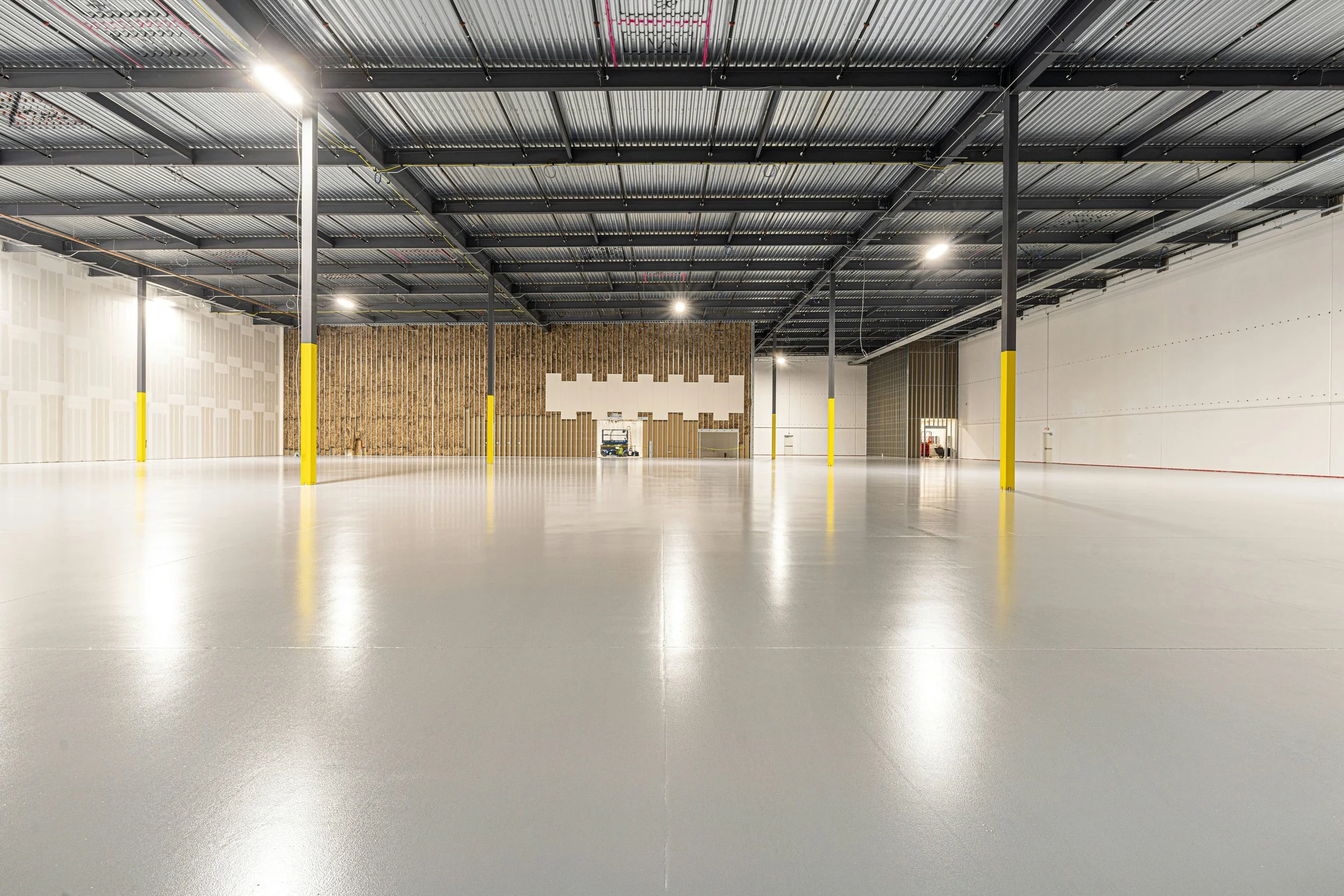 An empty indoor warehouse with a polished concrete floor, metallic ceiling, and yellow safety poles. The far wall has wooden insulation panels and a white section, with some equipment and a lift present.