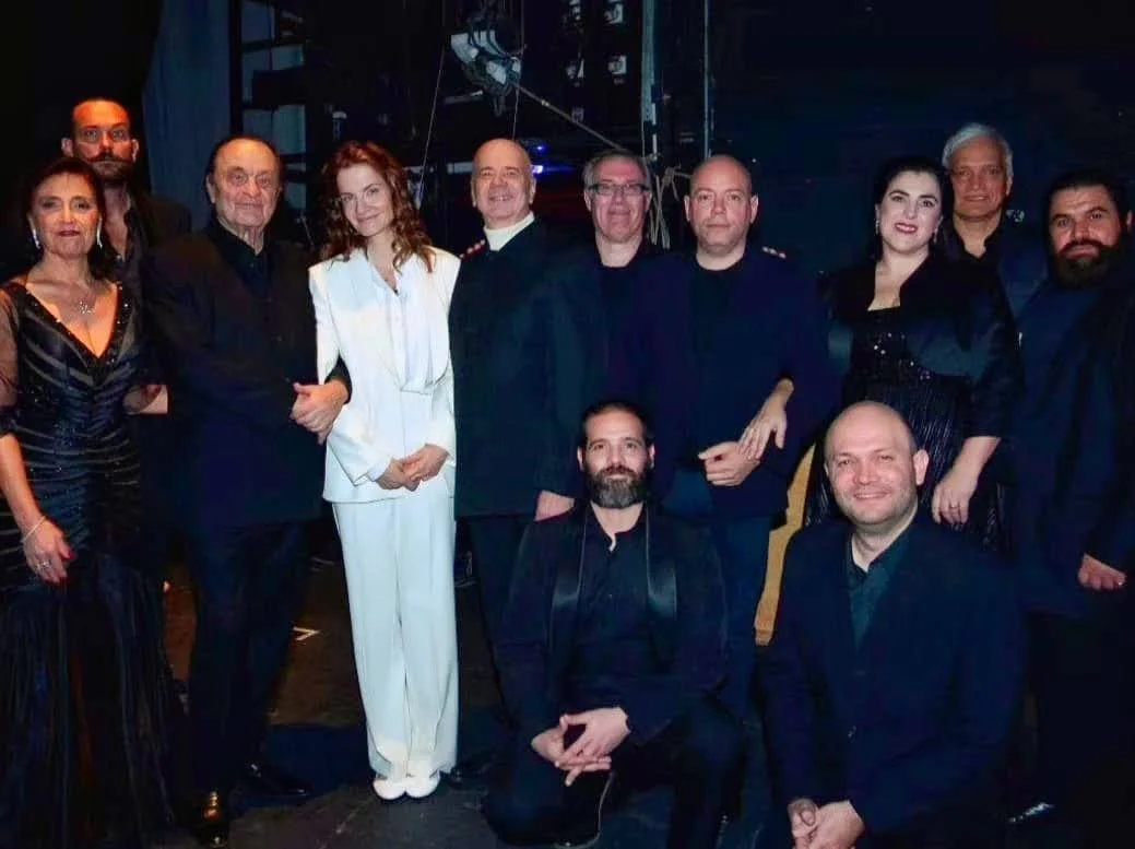 Grupo de personas posando para una foto en un escenario, algunos con vestimenta formal y otros con ropa negra, en un entorno oscuro con equipos de sonido visibles.