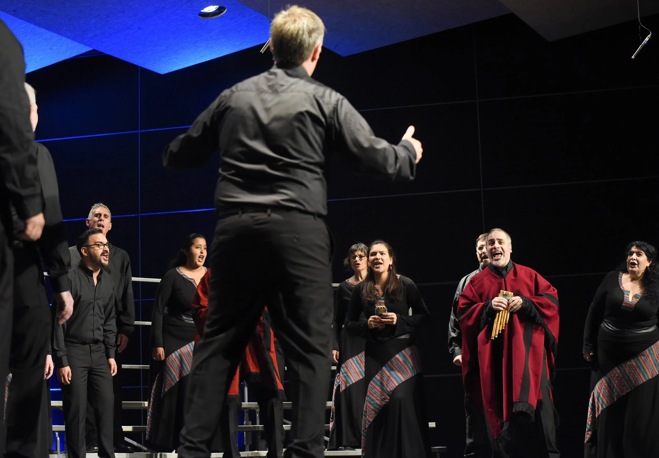 Un grupo de cantantes en un escenario, algunos con vestimenta tradicional, participando en una presentación musical con un director en primer plano.