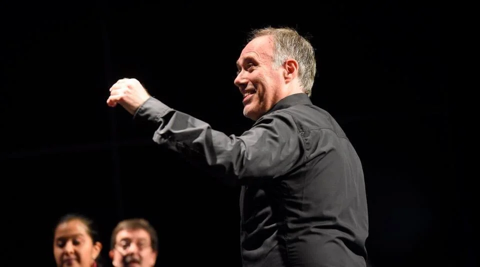 Hombre sonriendo y levantando la mano, con otras personas al fondo en un escenario oscuro.