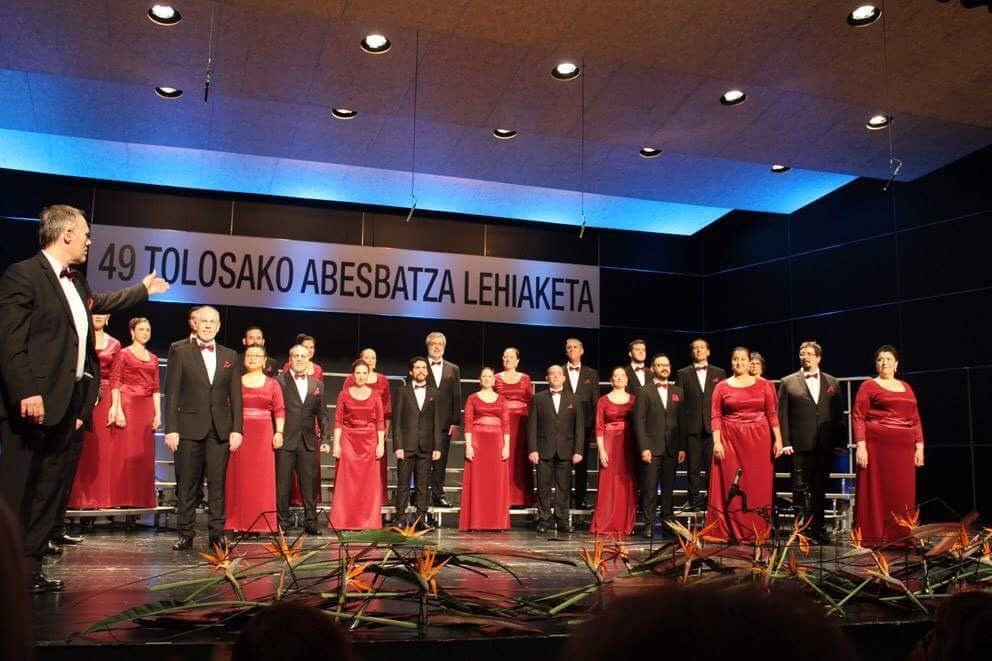 Un grupo de cantantes de coro en un escenario, vestidos con trajes formales negros y vestidos largos rojos, durante una presentación musical.