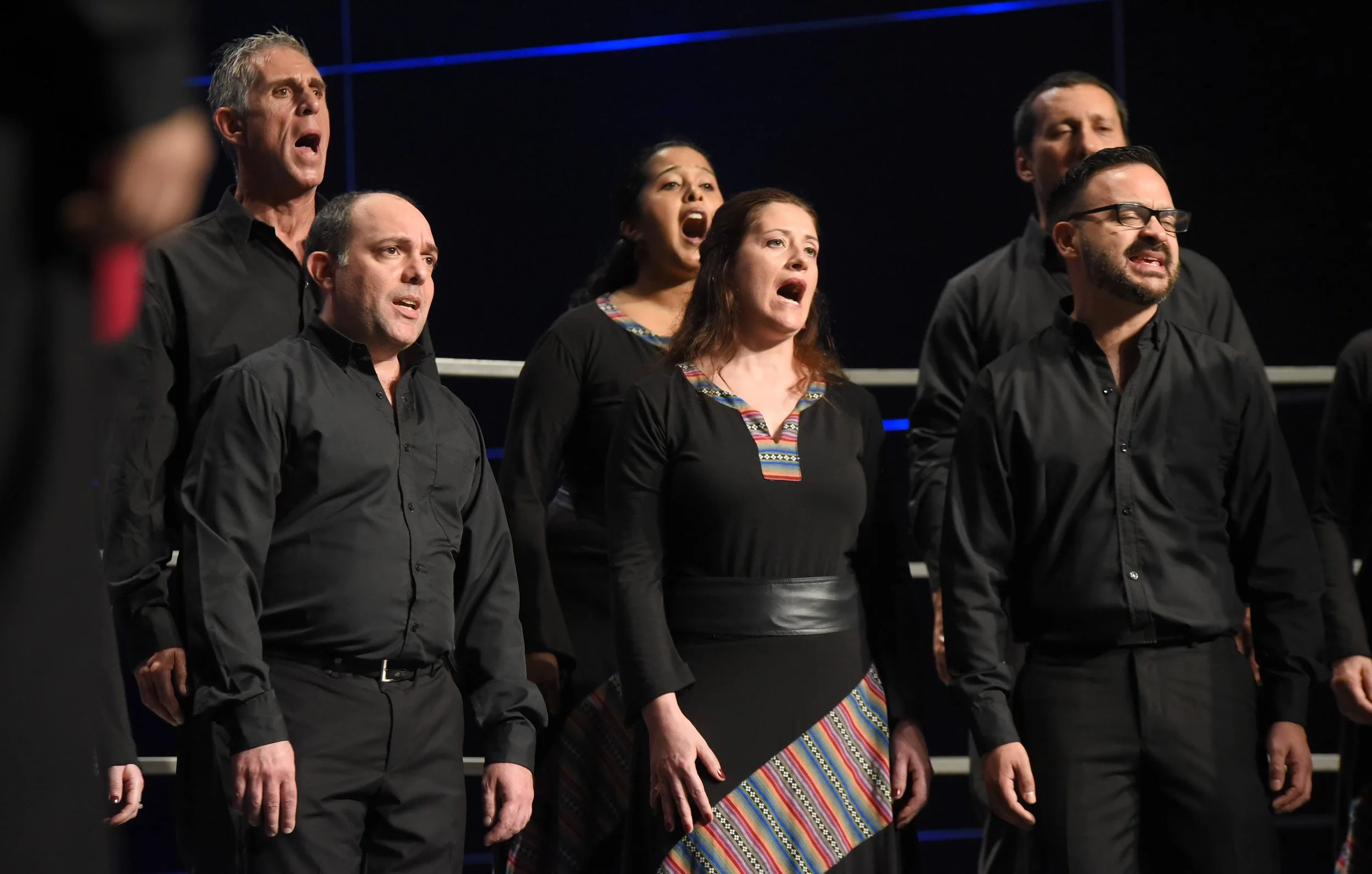 Grupo de personas cantando en un escenario, algunas con expresión intensa y emocional, vestidas con ropa negra, en un fondo oscuro con líneas azules.