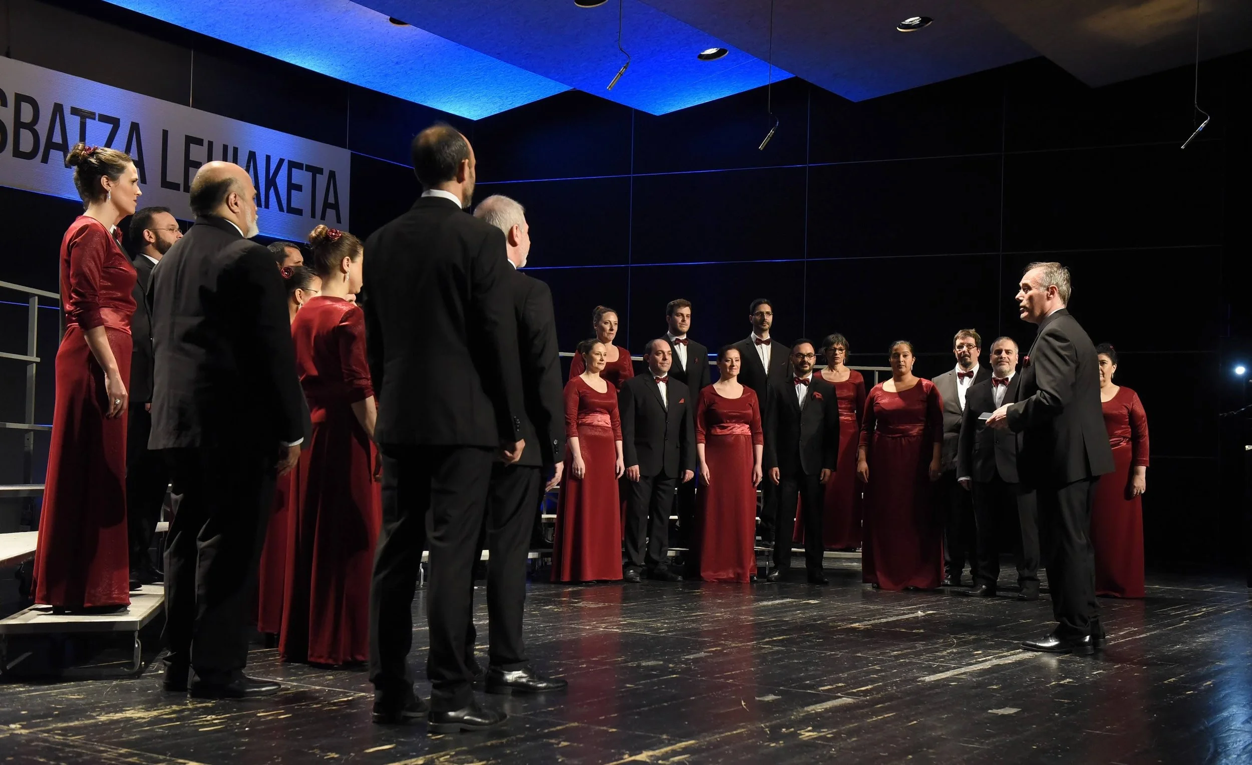 Un grupo de cantantes deópera en un escenario, vestidos con ropa formal, en un concierto en un teatro.