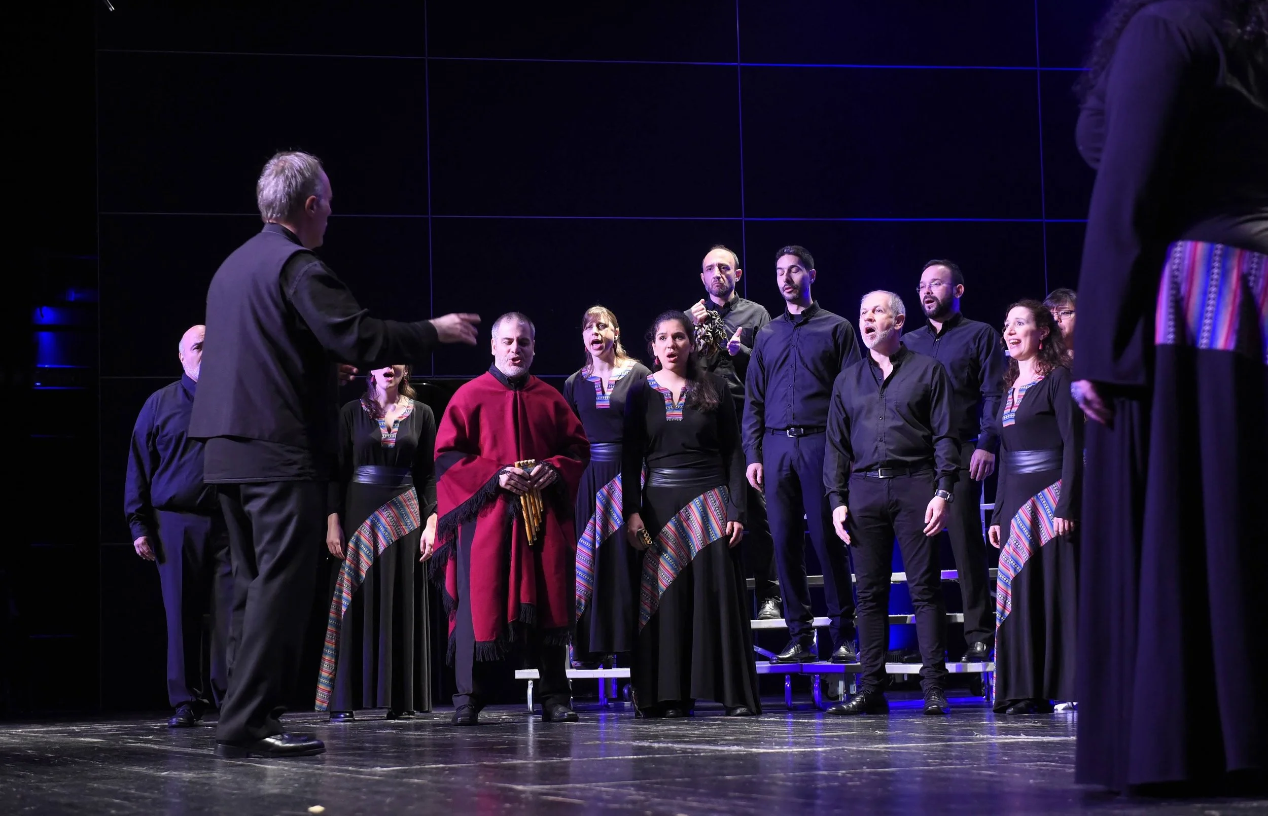 Un grupo de personas cantando en un escenario, un hombre con ropa negra parece estar dirigiendo el canto, algunas personas llevan trajes con detalles coloridos en las solapas o cinturillas.