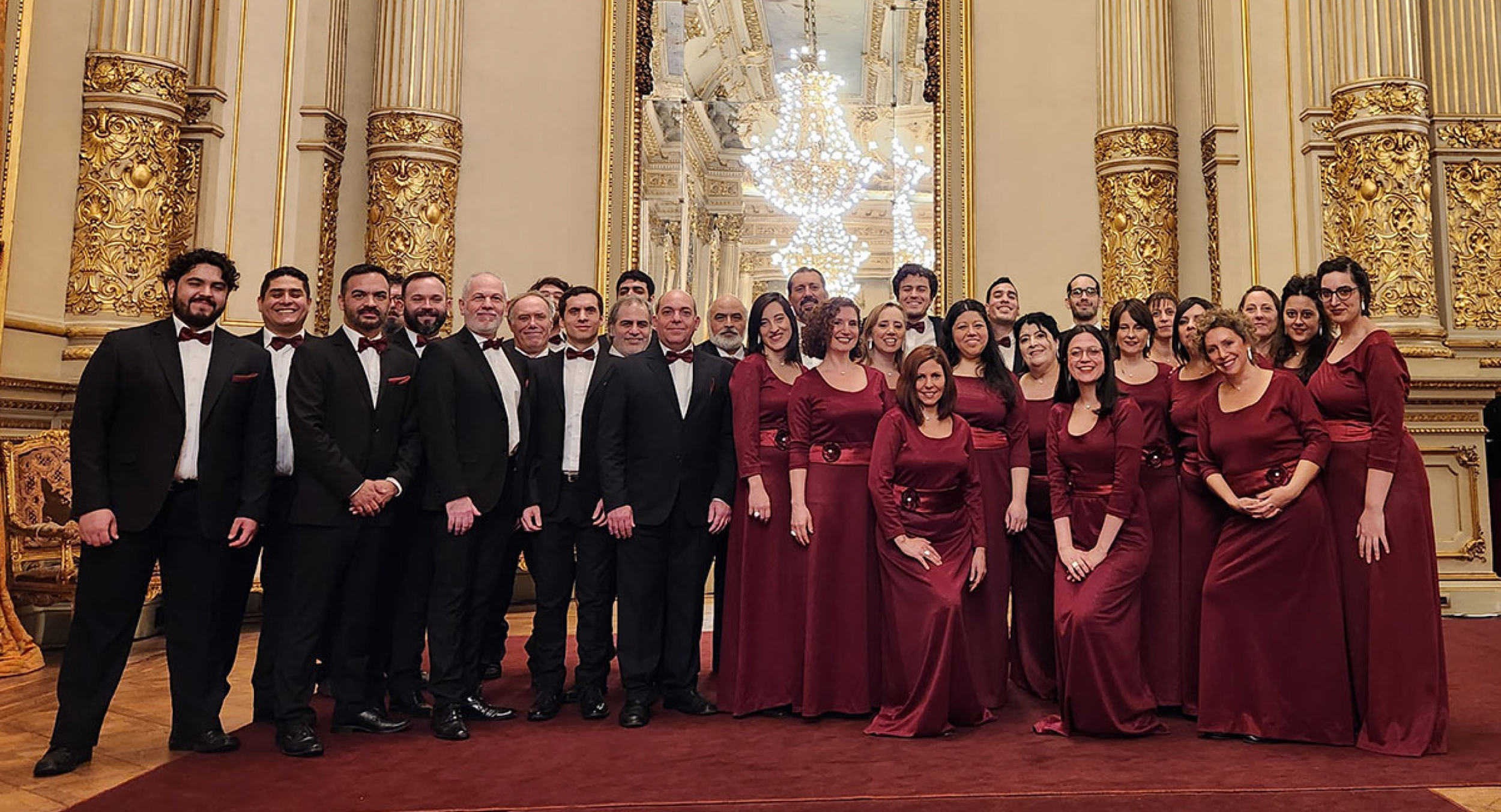 Grupo de personas vestidas con trajes formales en un salón elegante con adornos dorados y un gran espejo, posando para la foto.