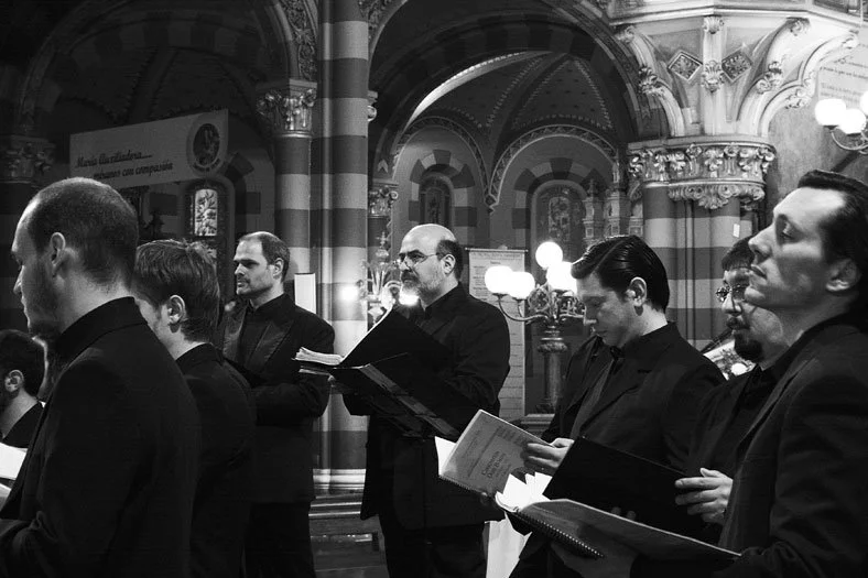 Imagen en blanco y negro de un grupo de hombres en una iglesia, probablemente coro, algunos con partituras en las manos, en un entorno con arquitectura religiosa.