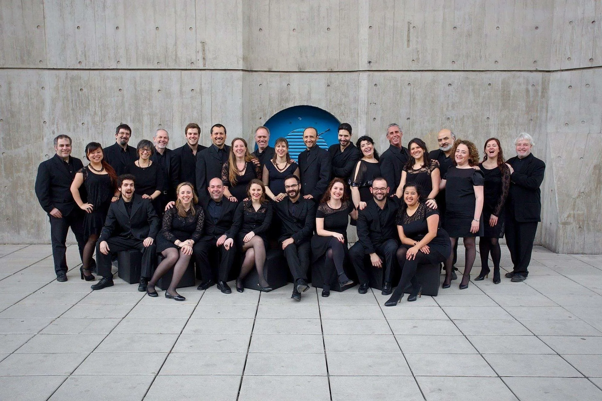 Grupo de personas vestidas formalmente en un ambiente exterior, posando para la fotografía frente a una pared de concreto con un círculo azul en el centro.