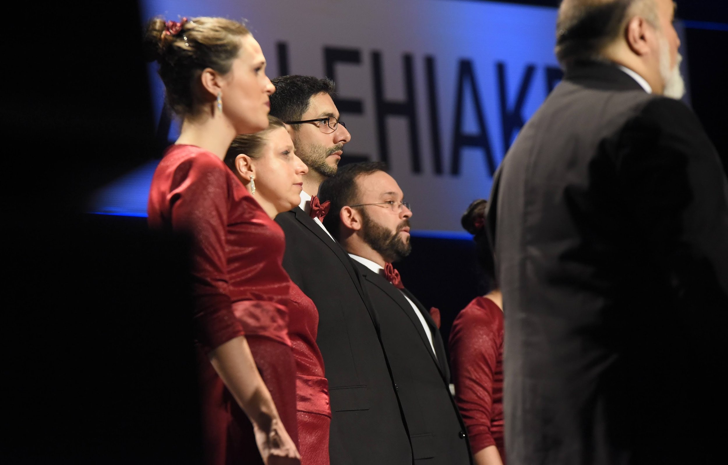 Grupo de personas en un evento formal, algunos con trajes elegantes, algunos con vestidos rojos, participando o asistiendo a una ceremonia o conferencia.