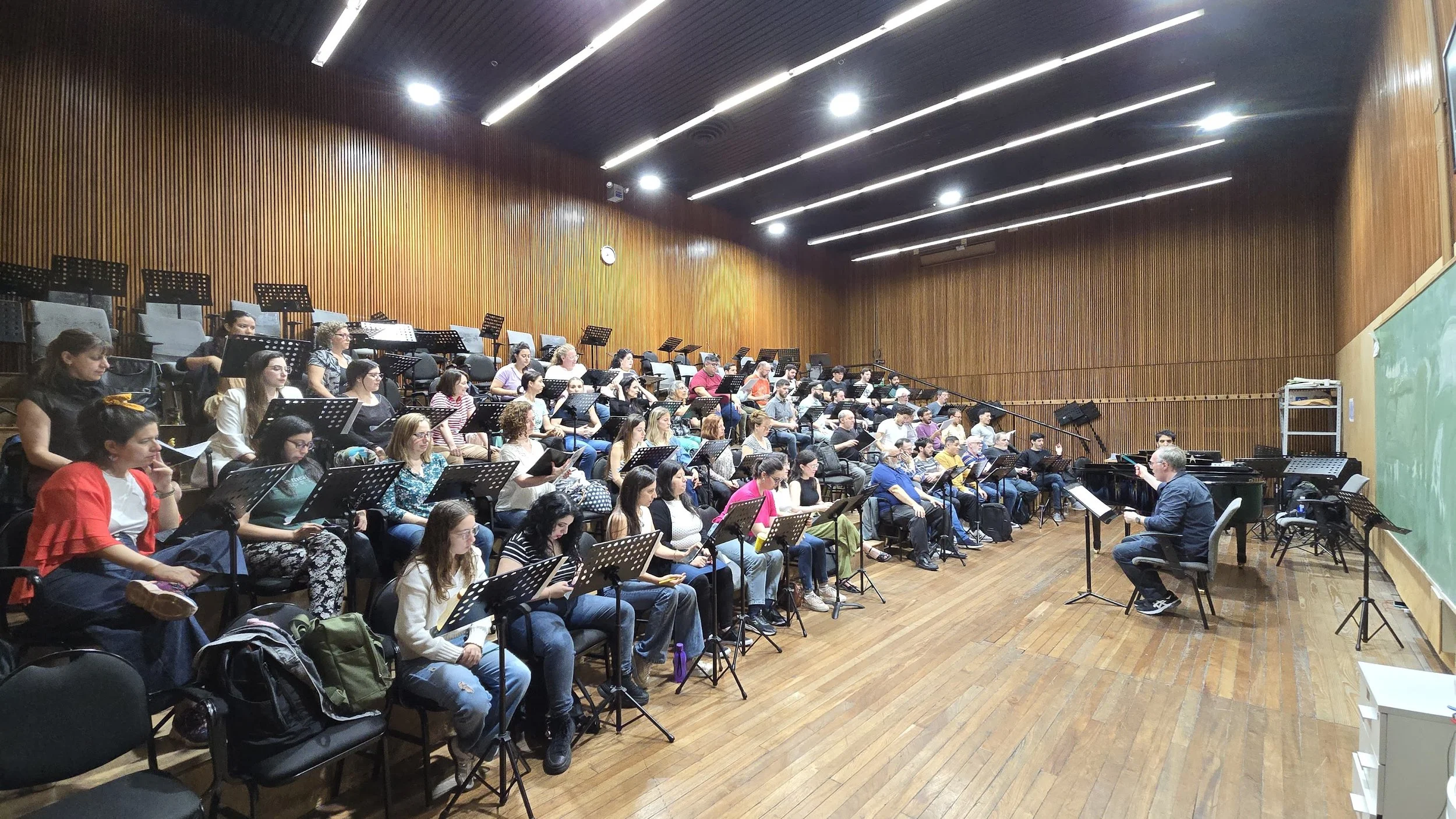 Un grupo grande de personas sentadas en un auditorio, escuchando a un director de orquesta que dirige con una mano, mientras tiene una partitura en el otro lado, en una sala con paredes de madera y un pizarrón verde a la derecha.