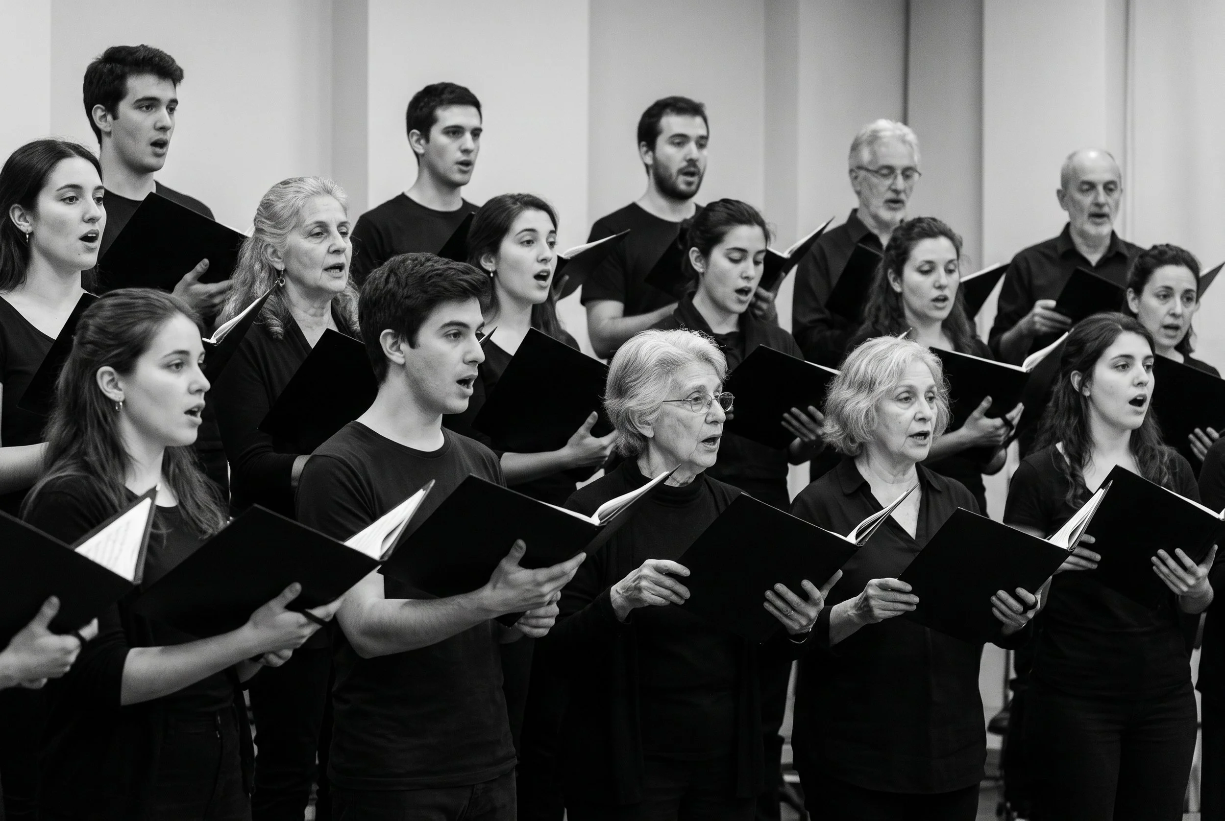Coro de personas de distintas edades cantando juntas en un ensayo, con carpetas de partitura en la mano.