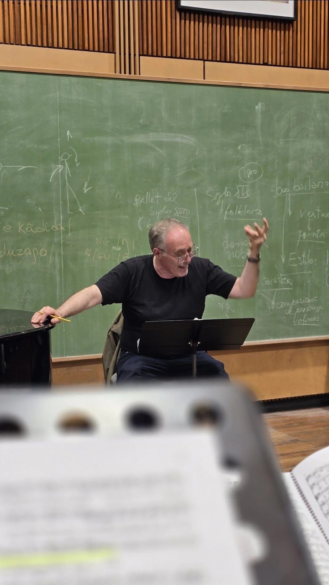 Un profesor de cabello gris y gafas, vestido con camiseta negra, enseñando en una pizarra verde cubierta de notas y diagramas, en un aula de clases.
