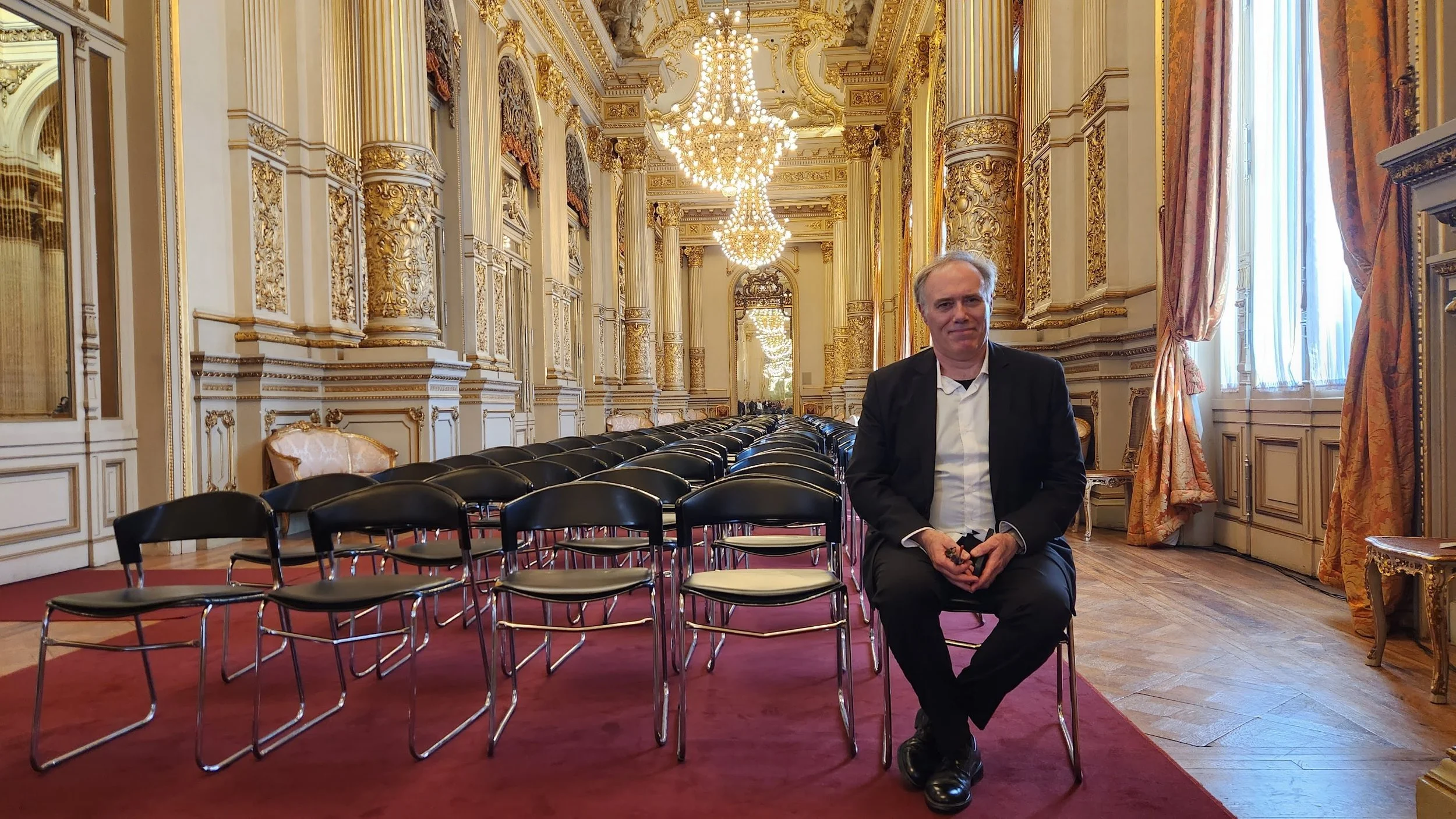 Hombre en un elegante salón con candelabros y cortinas doradas, sentado entre sillas negras, vestimenta formal.