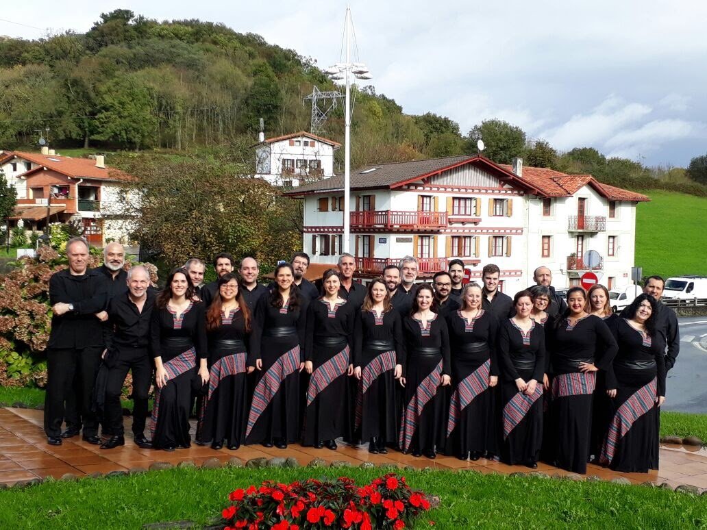 Grupo de personas vestidas con ropa negra y faldas con detalles coloridos, posando en un área exterior con casas y vegetación de fondo.