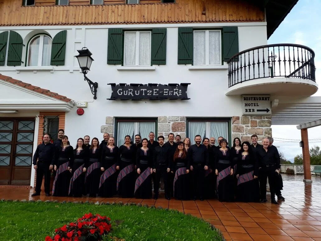 Grupo de personas con ropa tradicional frente a un hotel llamado Gukutze-Beri, en un edificio de estilo europeo con balcón de hierro y ventanas con persianas verdes.