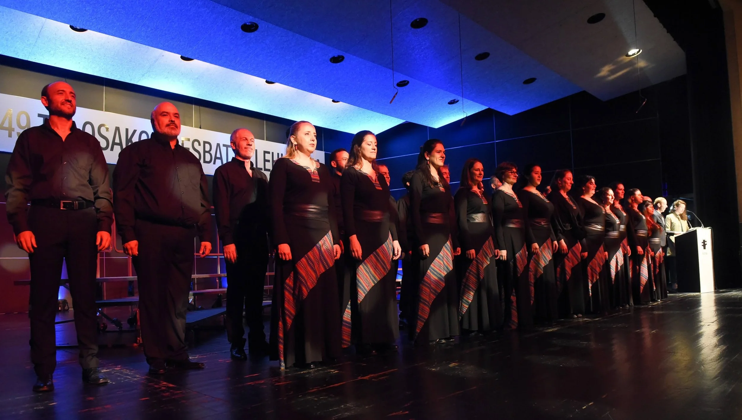 Grupo de personas en atuendos tradicionales en una ceremonia o evento en un escenario con iluminación azul y anuncios en el fondo.