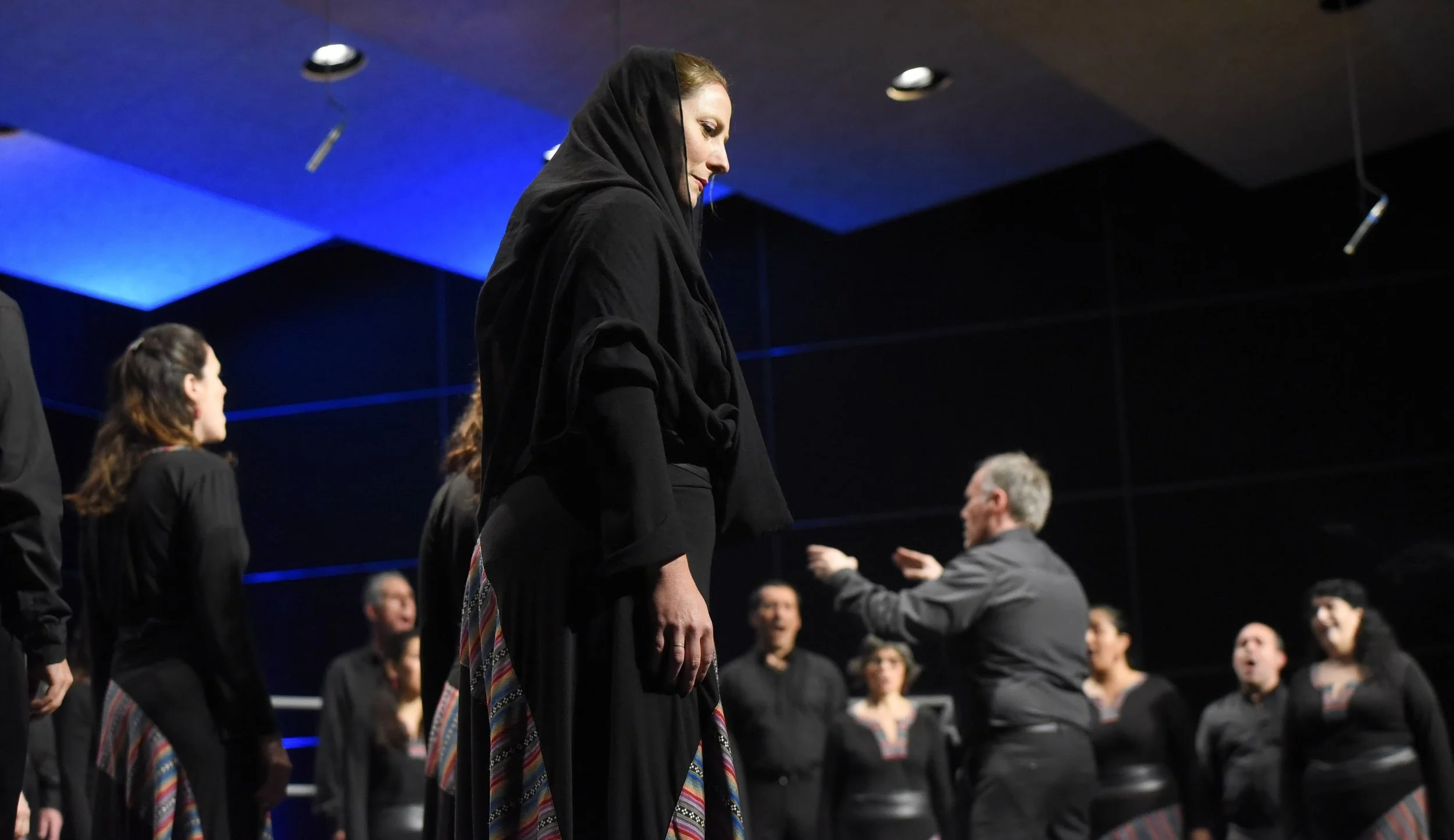 Mujer de cabello rubio y piel clara vestida con ropa negra, de perfil, en un entorno de concierto o ceremonia, rodeada de otras personas.