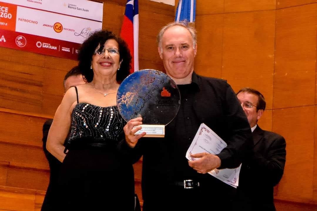 Un hombre y una mujer en un escenario de madera, levantando un premio en forma de globo terráqueo, con otras personas en el fondo y banderas a un lado.