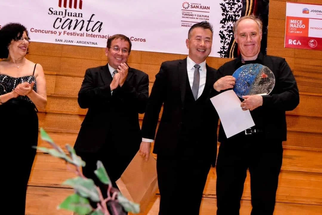 Personas en un escenario en un evento cultural en San Juan, Argentina, con un cartel que dice 'San Juan canta', algunos aplaudiendo y una persona en el centro recibiendo un premio o reconocimiento.